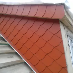 Fragment dachu z nowym, czerwonym pokryciem przypominającym łuskę, widoczne elementy konstrukcji dachu i okno.