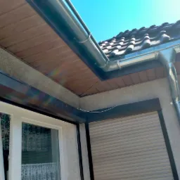 Nowe, srebrne rynny zamontowane na budynku z brązową podbitką dachową i ciemną dachówką, widok z dołu w słoneczny dzień.