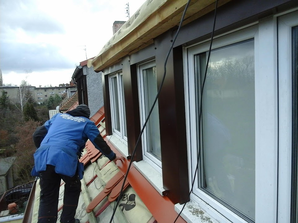 Pracownik w niebieskiej kurtce montuje brązowe obróbki blacharskie przy oknach dachowych na pomarańczowym dachu z dachówki ceramicznej, widoczne elementy izolacji i okablowanie.