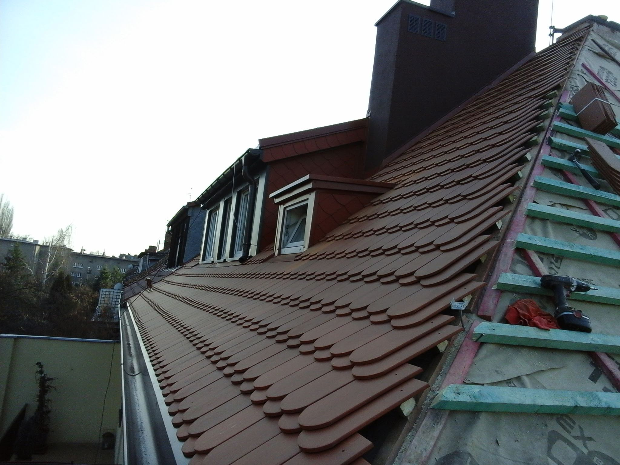 Ceglasty dach z dachówki bitumicznej w trakcie układania, widoczne łatki, narzędzia, komin w tle, panorama miejska w oddali.