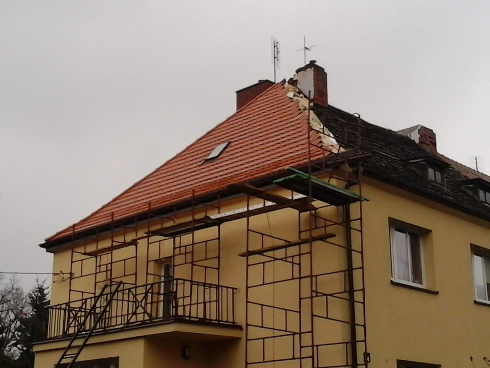Dwupiętrowy budynek z częściowo odnowionym dachem z czerwonej dachówki, rusztowanie przy ścianie, część dachu w trakcie wymiany, widoczne stare poszycie dachu przy kominie.