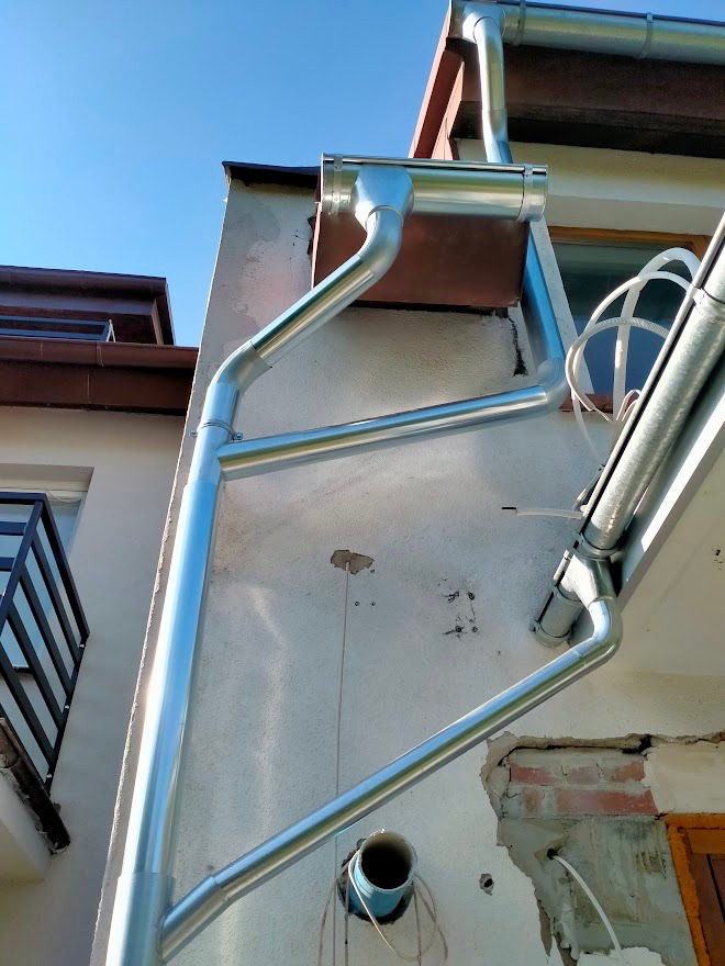 Nowe, błyszczące, srebrne rynny spustowe zamontowane na elewacji budynku z widocznymi śladami wcześniejszych prac i wystającymi przewodami.