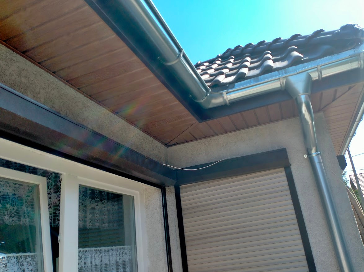 Nowe, srebrne rynny zamontowane na budynku z brązową podbitką dachową i ciemną dachówką, widok z dołu w słoneczny dzień.