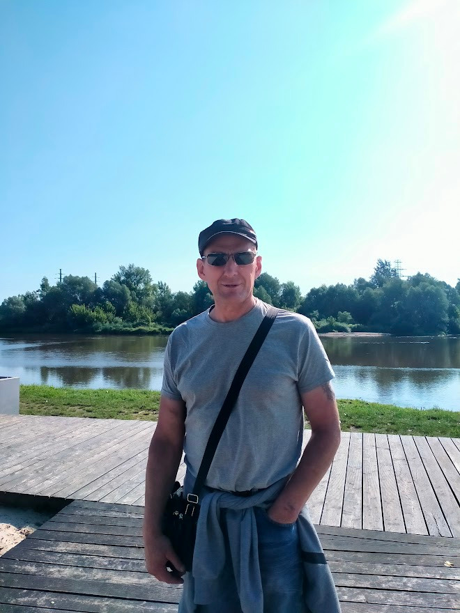 Mężczyzna w okularach przeciwsłonecznych i czapce z daszkiem, stojący na drewnianym pomoście nad rzeką w słoneczny dzień. W tle zieleń drzew i błękitne niebo.