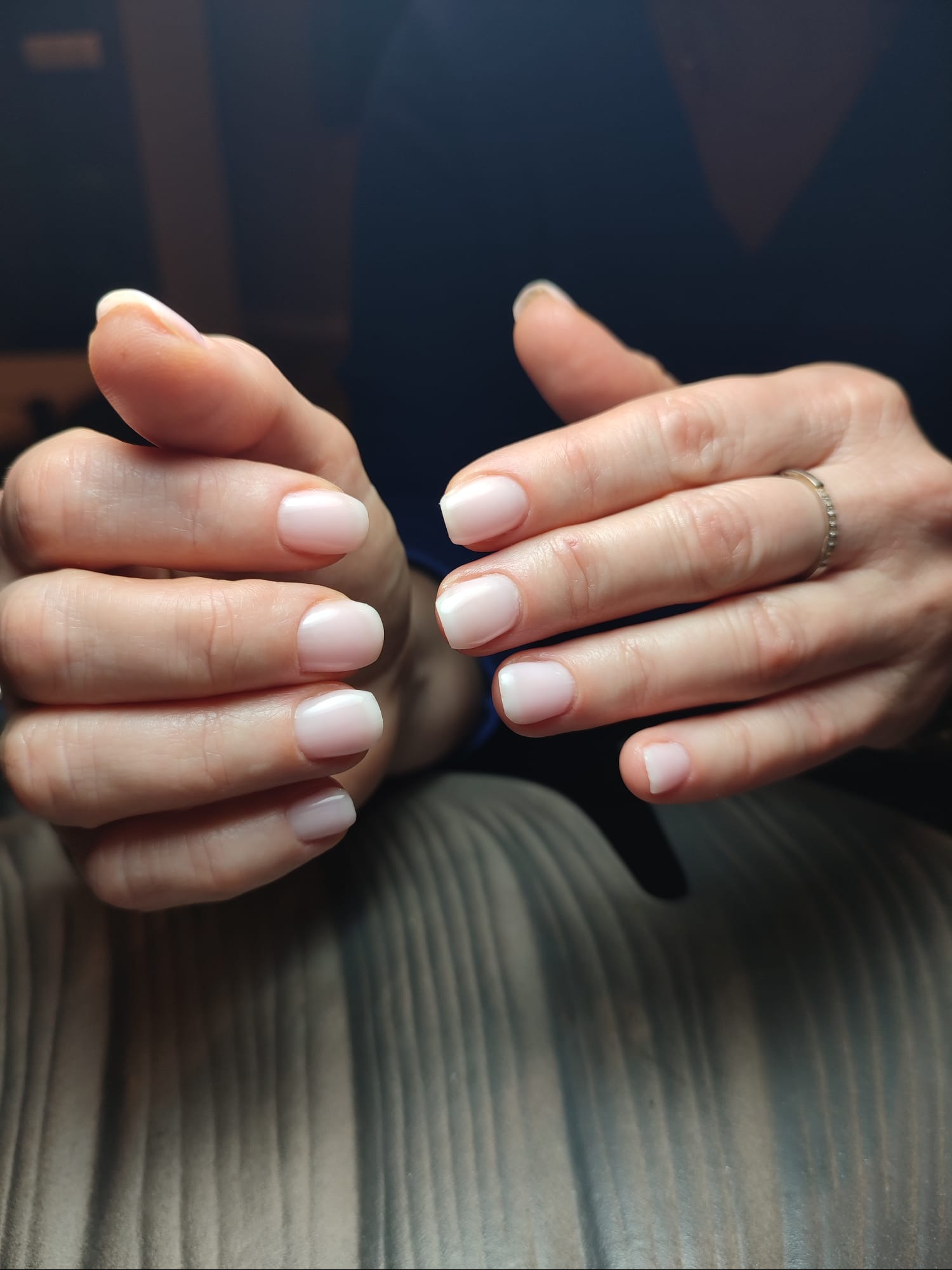 Dłonie z delikatnym, mlecznym manicure hybrydowym o kwadratowym kształcie, pierścionek na palcu serdecznym prawej dłoni, tło w odcieniach szarości.