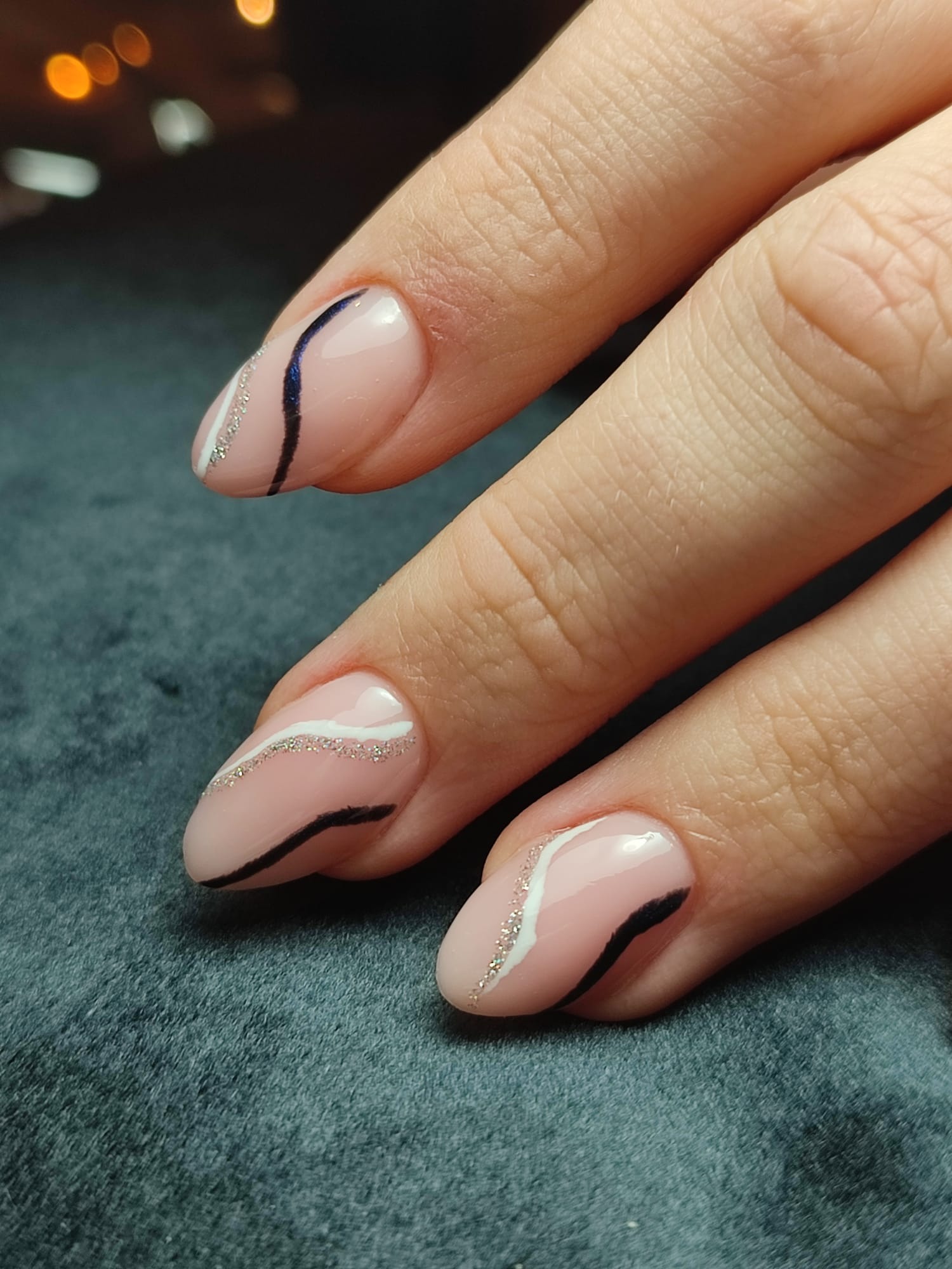 Dłoń z trzema paznokciami w odcieniach nude, ozdobionymi asymetrycznymi wzorami w kolorach czarnym, białym i srebrnym brokatem, na tle ciemnego, aksamitnego materiału.