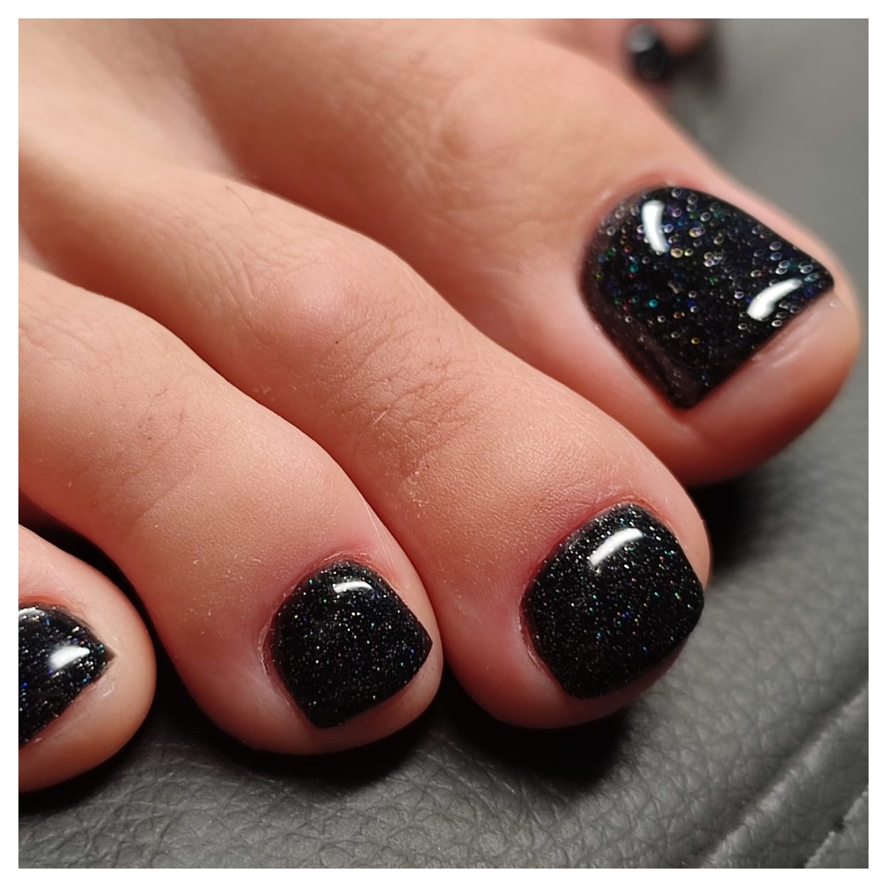 Zbliżenie na zadbane stopy z czarnym, brokatowym lakierem hybrydowym na paznokciach, widoczne drobne, wielokolorowe refleksy.