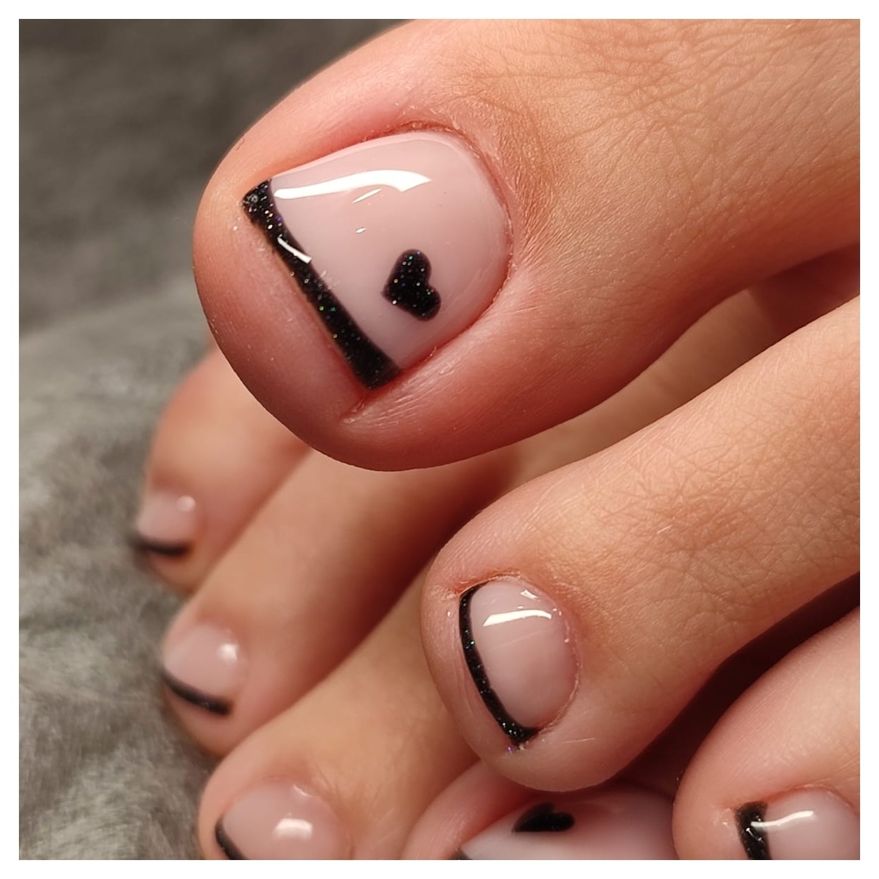 Zbliżenie na pomalowane paznokcie u stóp z jasnoróżowym lakierem, czarnym paskiem po przekątnej i małym czarnym serduszkiem.