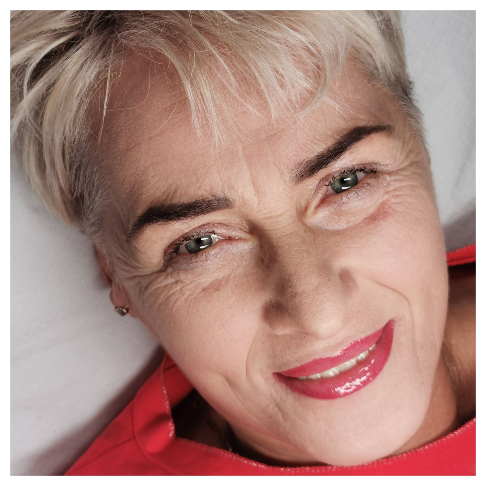 Kobieta o krótkich blond włosach i zielonych oczach z pełnym, błyszczącym makijażem ust w odcieniu fuksji, leżąca na białym tle, uśmiechnięta.