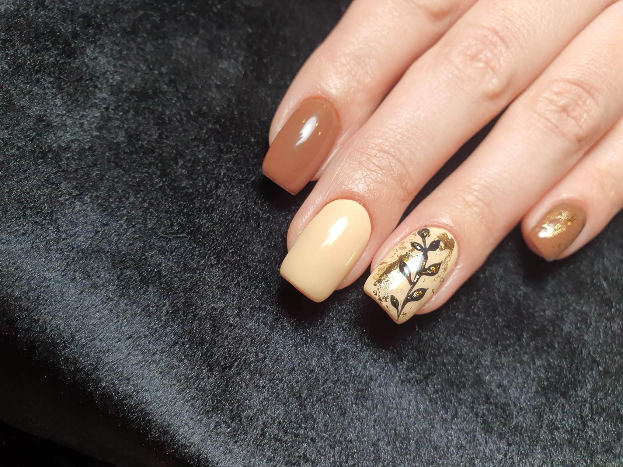 Dłoń z pomalowanymi paznokciami w odcieniach beżu, brązu i złota, jeden paznokieć zdobiony motywem roślinnym na ciemnym, aksamitnym tle.