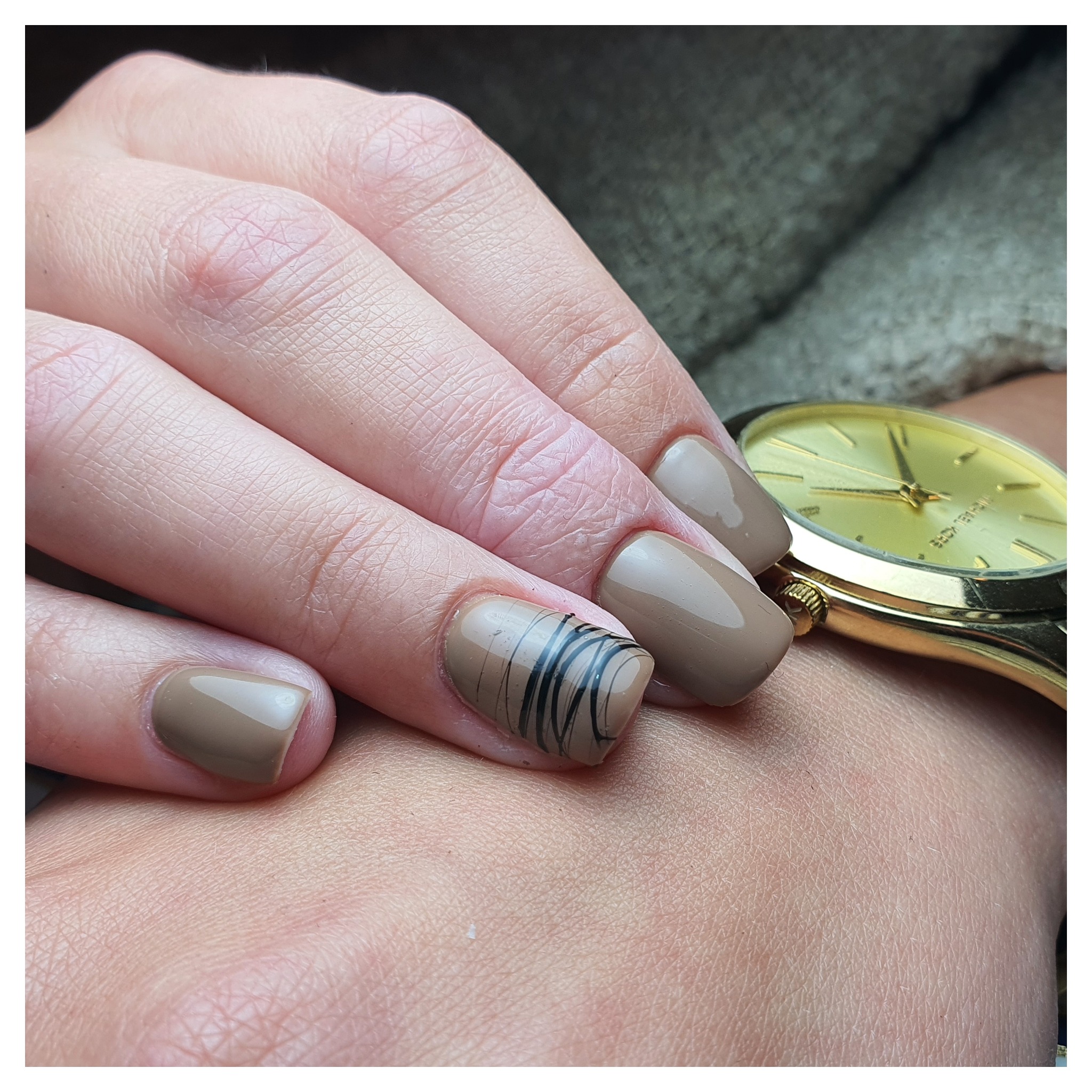 Paznokcie w odcieniach brązu, jeden z czarnymi liniami, na tle złotej bransoletki zegarka.