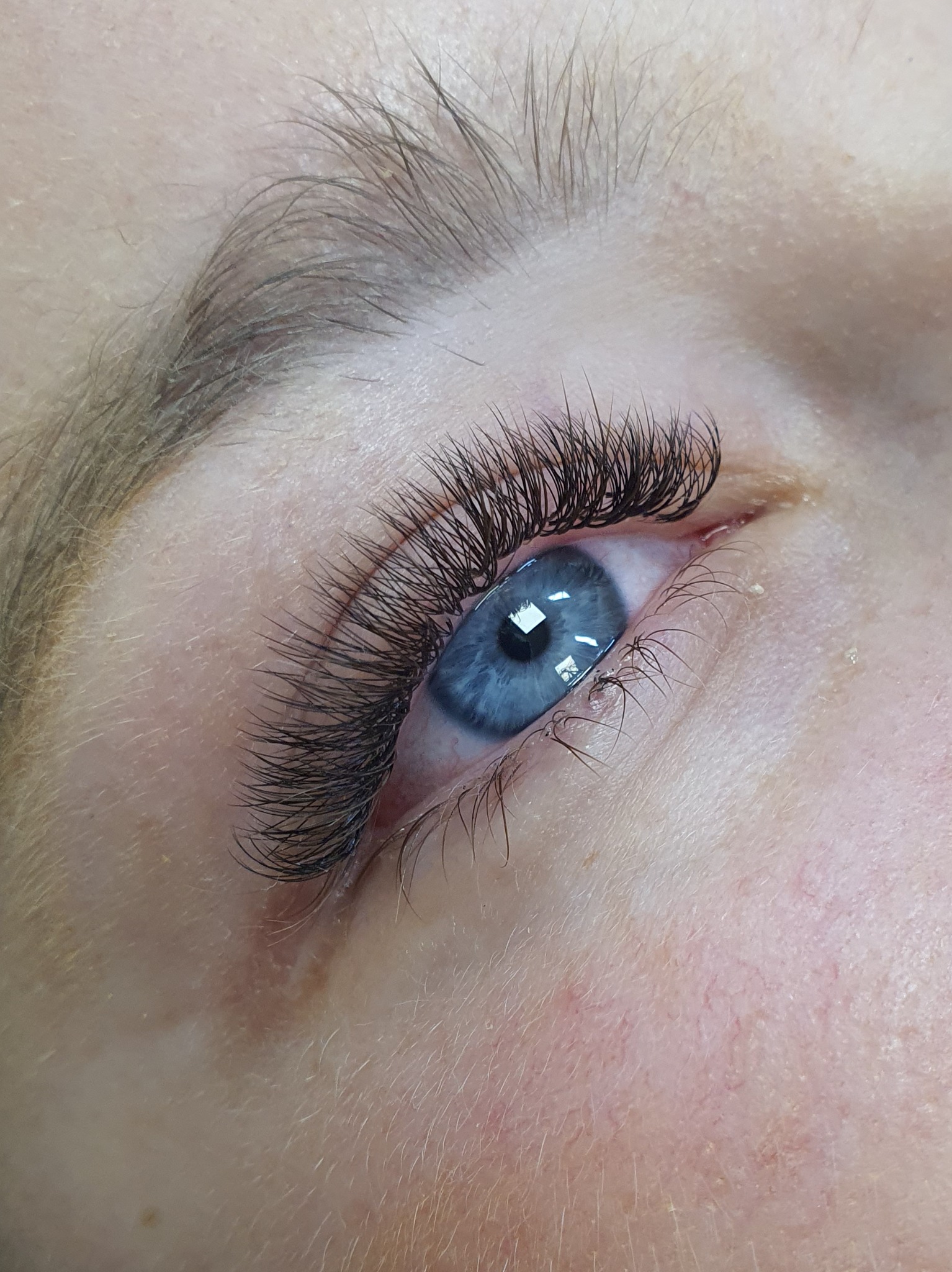 Zbliżenie na oko z długimi, gęstymi, czarnymi rzęsami, podkreślającymi intensywny, błękitny kolor tęczówki. Widoczny delikatny makijaż oka i naturalny odcień skóry.