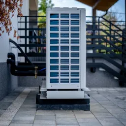 Biała jednostka zewnętrzna z widocznymi niebieskimi lamelami, zamontowana na czarnym cokole przy budynku z czarnymi rurami i schodami z metalową balustradą w tle.