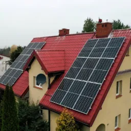 Dom jednorodzinny z czerwonym dachem i panelami słonecznymi na połaciach, widok z góry na tle zieleni.