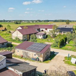 MR Energy Spółka z Ograniczoną Odpowiedzialnością - Widok z góry na wiejski krajobraz z zabudowaniami gospodarczymi, z których dwa mają zainstalowane panele fotowoltaiczne na dachu, w tle pola uprawne i las