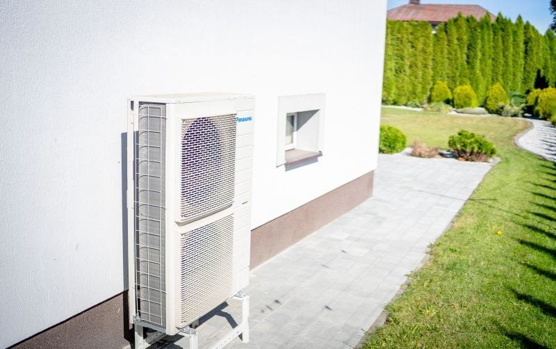 Zewnętrzna jednostka klimatyzacji Panasonic zamontowana na wspornikach przy białej ścianie budynku z widocznym oknem i zadbanym ogrodem w tle.