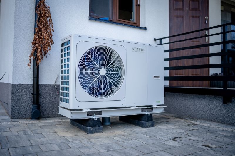 Biała pompa ciepła marki Netsu zamontowana na szarej kostce brukowej przed budynkiem z brązowymi drzwiami i oknem, widoczny czarny spust rynnowy i uschnięte pnącze.