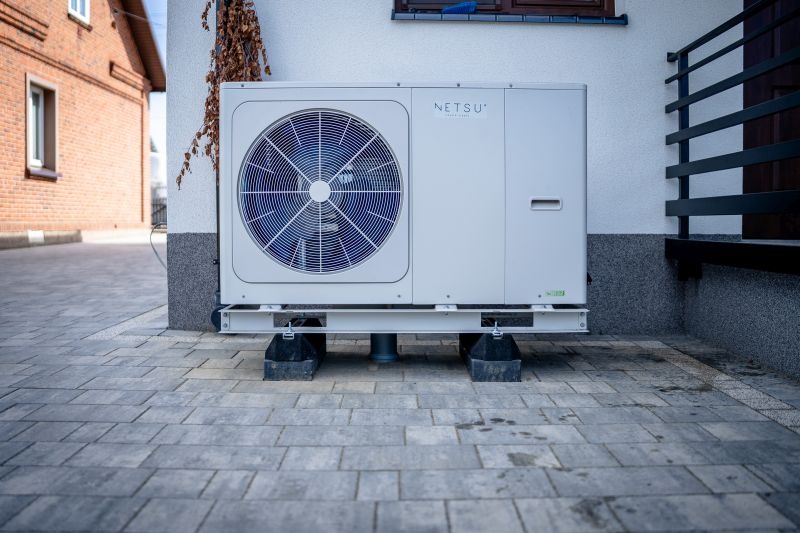 Biała jednostka zewnętrzna z wentylatorem marki Netsu zamontowana na metalowej ramie i gumowych podkładkach, ustawiona na szarej kostce brukowej przy ścianie budynku.