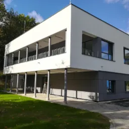 Nowoczesny, dwukondygnacyjny budynek biurowy z płaskim dachem, widoczny z zewnątrz w Koszalinie, z szarymi panelami elewacyjnymi i białymi ścianami, wsparty na kolumnach, z zielonym trawnikiem...