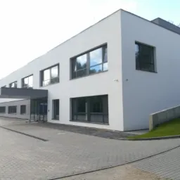 Nowoczesny, dwukondygnacyjny budynek biurowy z białymi ścianami i dużymi oknami, widok z zewnątrz, z szarym chodnikiem z kostki brukowej.