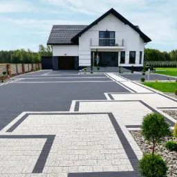 Nowoczesny dom jednorodzinny z podjazdem wyłożonym dwukolorową kostką brukową w geometryczne wzory, otoczony zadbanym trawnikiem i ozdobnymi drzewkami.