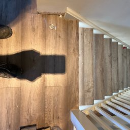 Ar-Wi - Montaż paneli podłogowych imitujących drewno na schodach w Szczecinie, widok z góry na fragment podłogi i schody z zamontowanymi białymi barierkami, w tle widoczne narzędzia i buty osoby montującej.