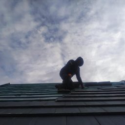 Ar-Wi - Monter w ciemnej odzieży roboczej układa ciemne dachówki na dachu, widoczne fragmenty hydroizolacji i pochmurne niebo w tle.