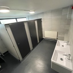 Ar-Wi - Wnętrze publicznej toalety po remoncie w Szczecinie: czarne kabiny WC z białymi ściankami działowymi, biała umywalka z dwoma kranami oraz grzejnik na tle szarych płytek ściennych.