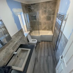 Ar-Wi - Wnętrze łazienki w trakcie remontu, widoczne fragmenty ścian z naniesioną farbą, szare płytki na ścianach i podłodze imitujące drewno, umywalka z czarnym brokatowym blatem, toaleta oraz grzejnik...