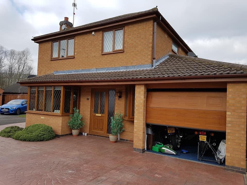 Dwupiętrowy dom z brązowej cegły z garażem, brązowymi oknami z kratami i częściowo otwartą bramą garażową, ukazującą motocykle w środku, oraz brukowanym podjazdem.