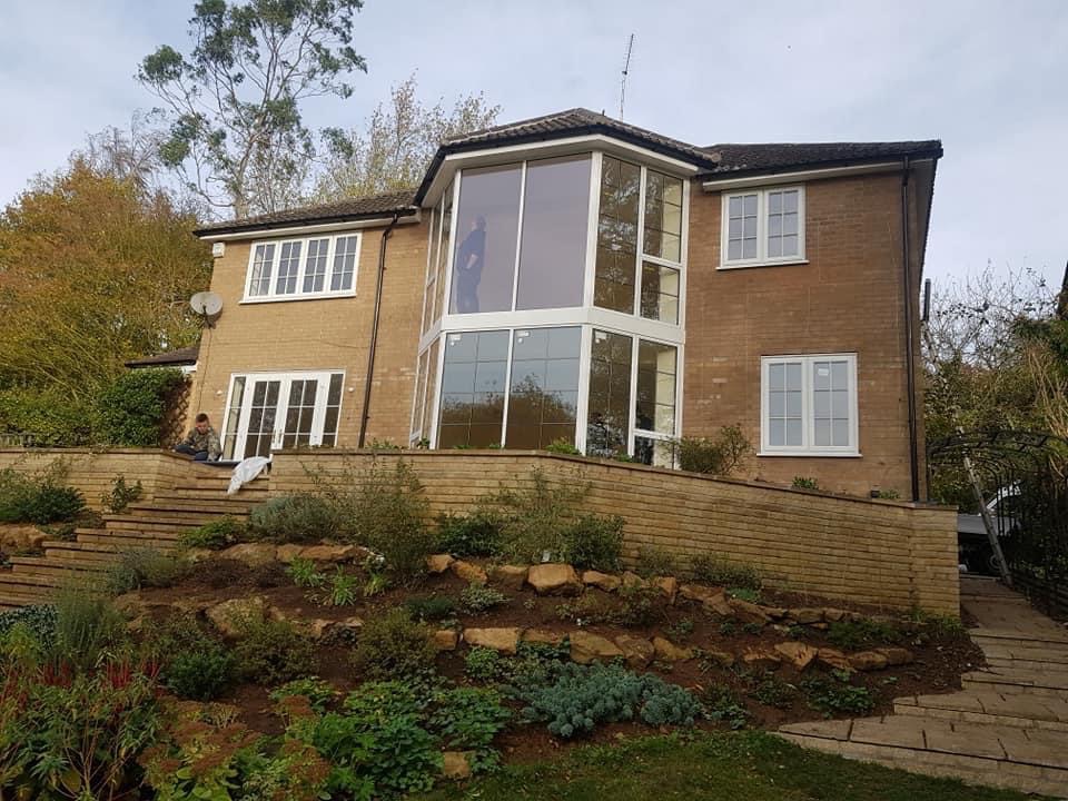 Dom z cegły z nową, przeszkloną dobudówką, widok z ogrodu z tarasami i roślinnością.