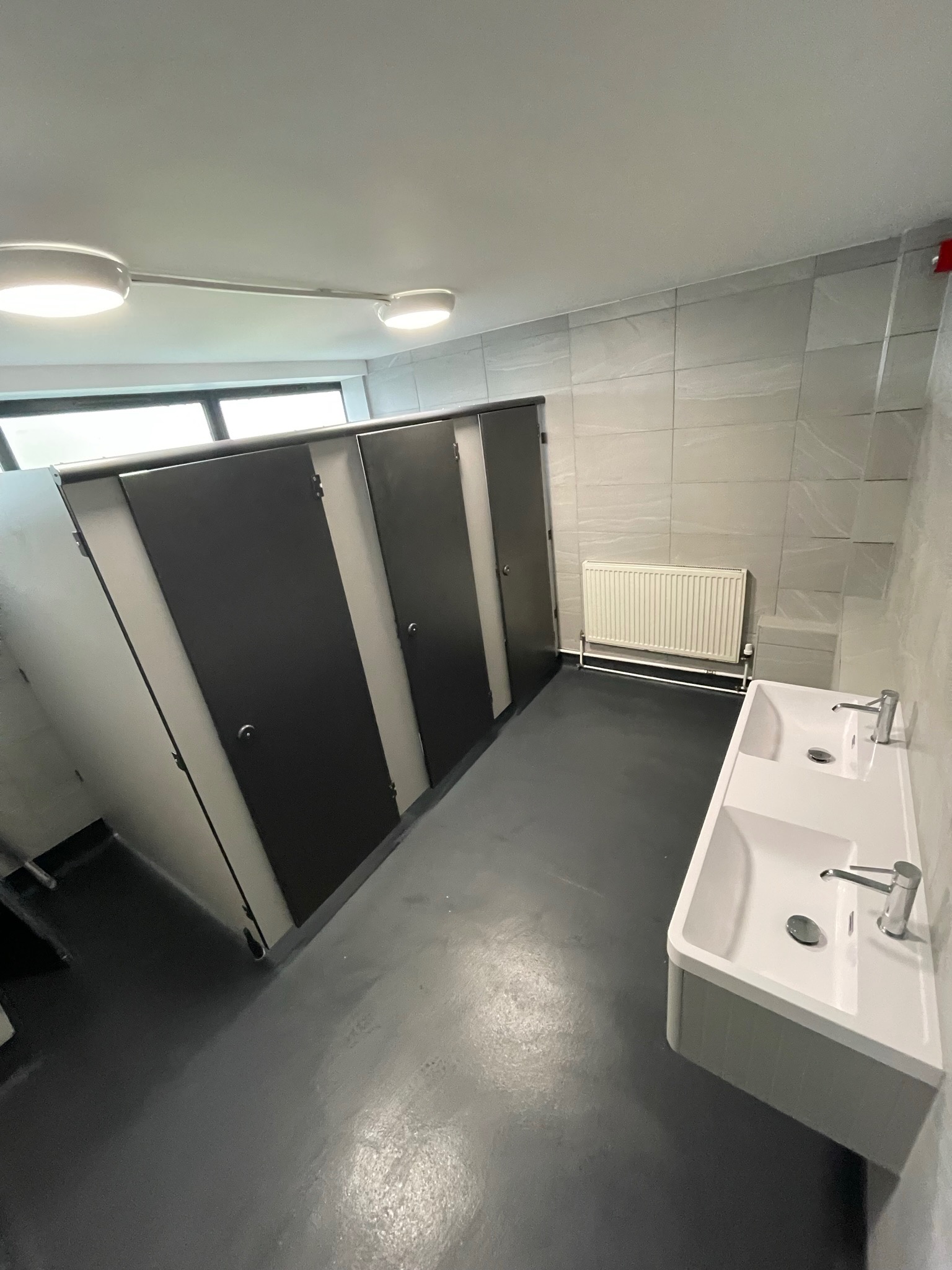 Wnętrze publicznej toalety z kabinami WC o czarnych drzwiach, dwukomorową białą umywalką z chromowanymi bateriami, szarymi płytkami ściennymi i grzejnikiem.