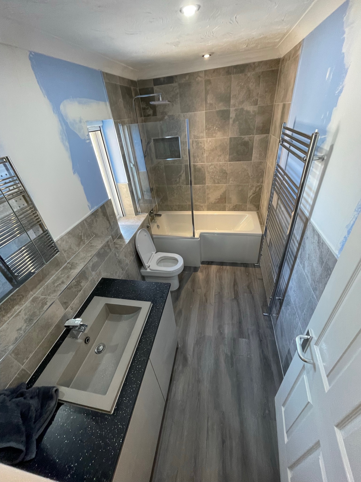 Wnętrze łazienki w trakcie remontu, widoczne fragmenty niedomalowanych ścian, nowe płytki, wanna z kabiną prysznicową, umywalka z czarnym blatem i grzejnik drabinkowy.