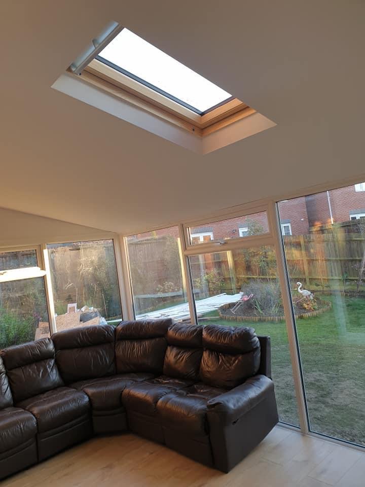Wnętrze jasnego pokoju z dużym, skórzanym narożnikiem, widokiem na ogród przez okna i oknem dachowym w suficie, podkreślającym doświetlenie pomieszczenia.