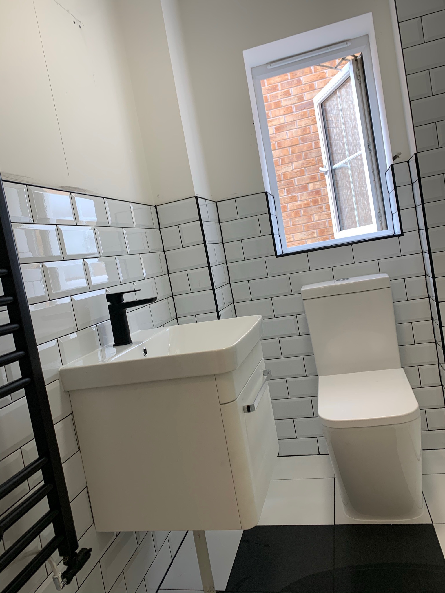 Nowoczesna łazienka z białą armaturą, czarną baterią umywalkową i grzejnikiem drabinkowym, wykończona białymi płytkami typu metro z czarną fugą oraz czarno-białą podłogą, okno otwarte.