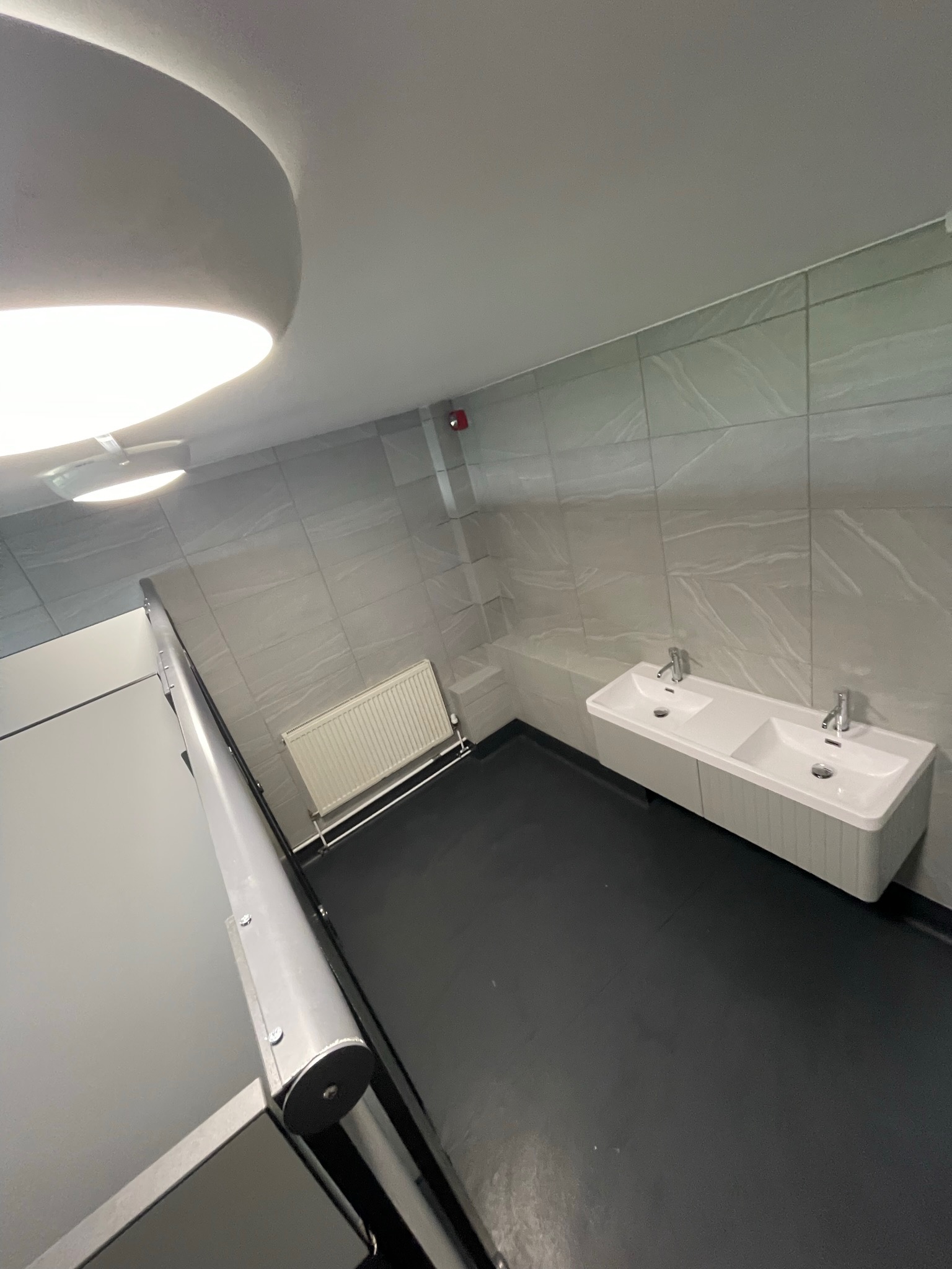 Nowoczesna łazienka z dwoma umywalkami nablatowymi i grzejnikiem panelowym, wykończona płytkami imitującymi marmur, oświetlona lampą sufitową.