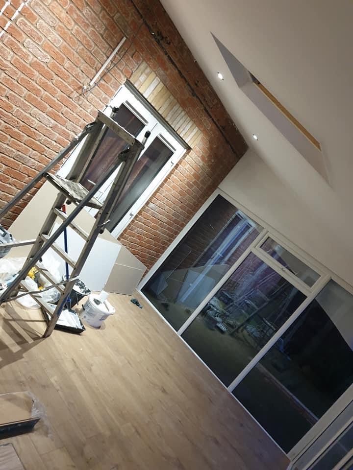 Wnętrze w trakcie prac wykończeniowych: widoczna ściana z cegły, okno z białymi ramami, drewniana podłoga, drabina i narzędzia malarskie.