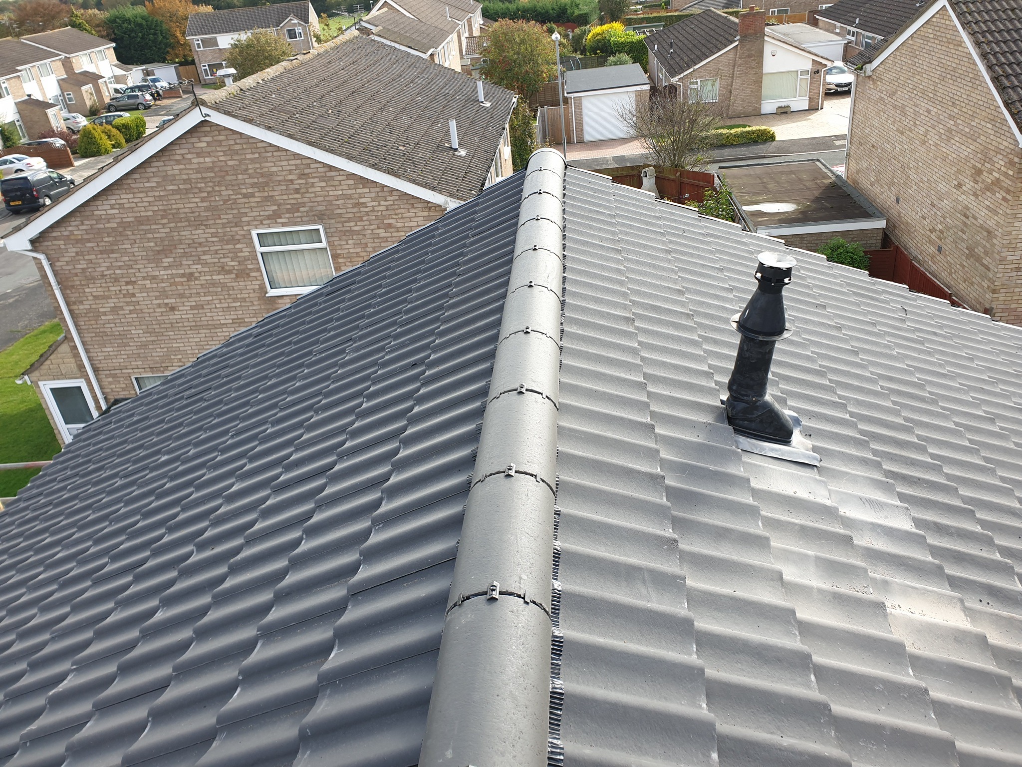 Widok z góry na nowy, szary dach z ceramicznej dachówki z centralnym kominem wentylacyjnym i systemem wentylacji kalenicy, otoczony domami jednorodzinnymi.