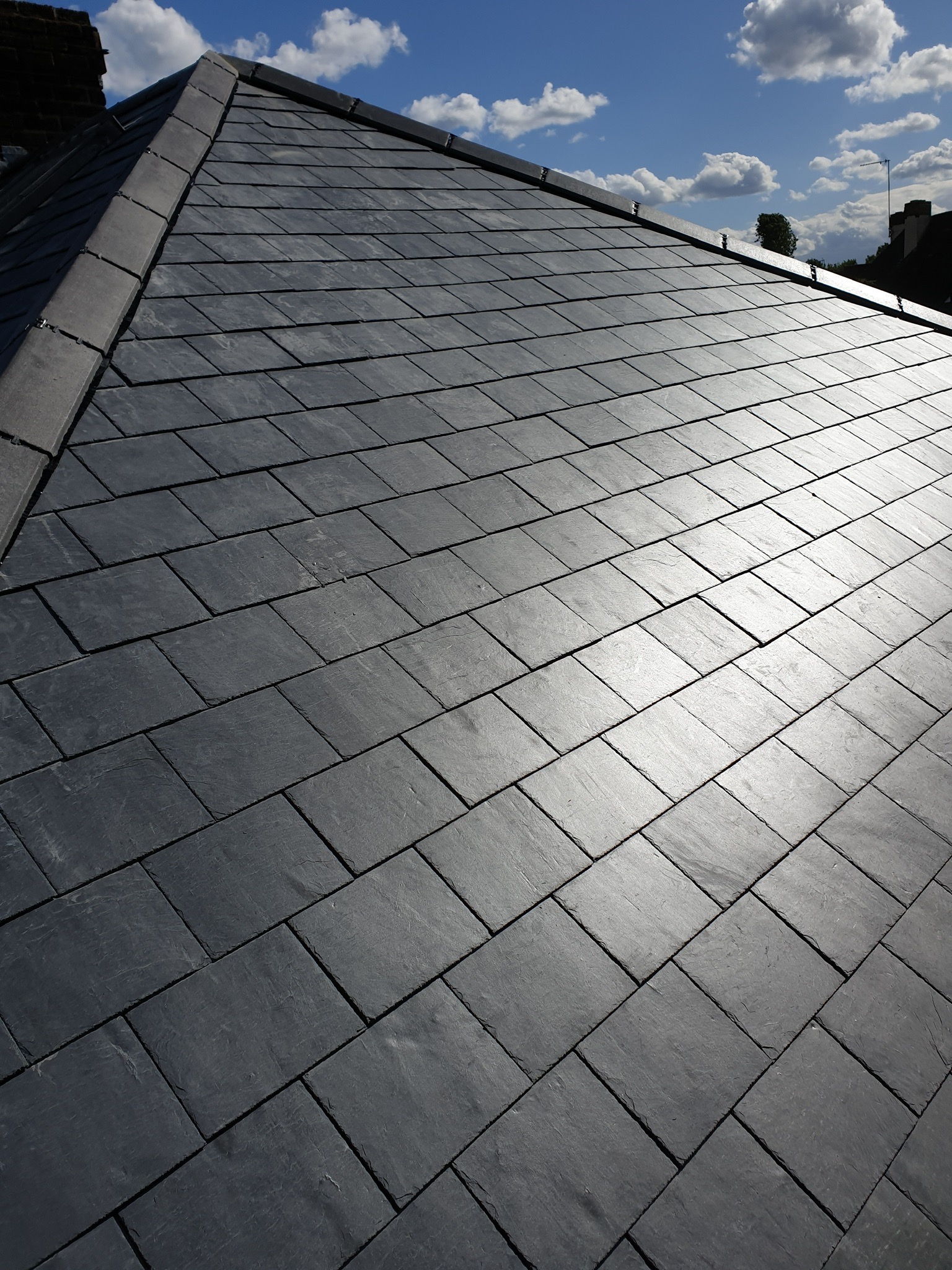 Nowy dach pokryty ciemnoszarym, łupkowym materiałem, widoczny z bliska pod kątem, z fragmentem komina i niebieskim niebem w tle.