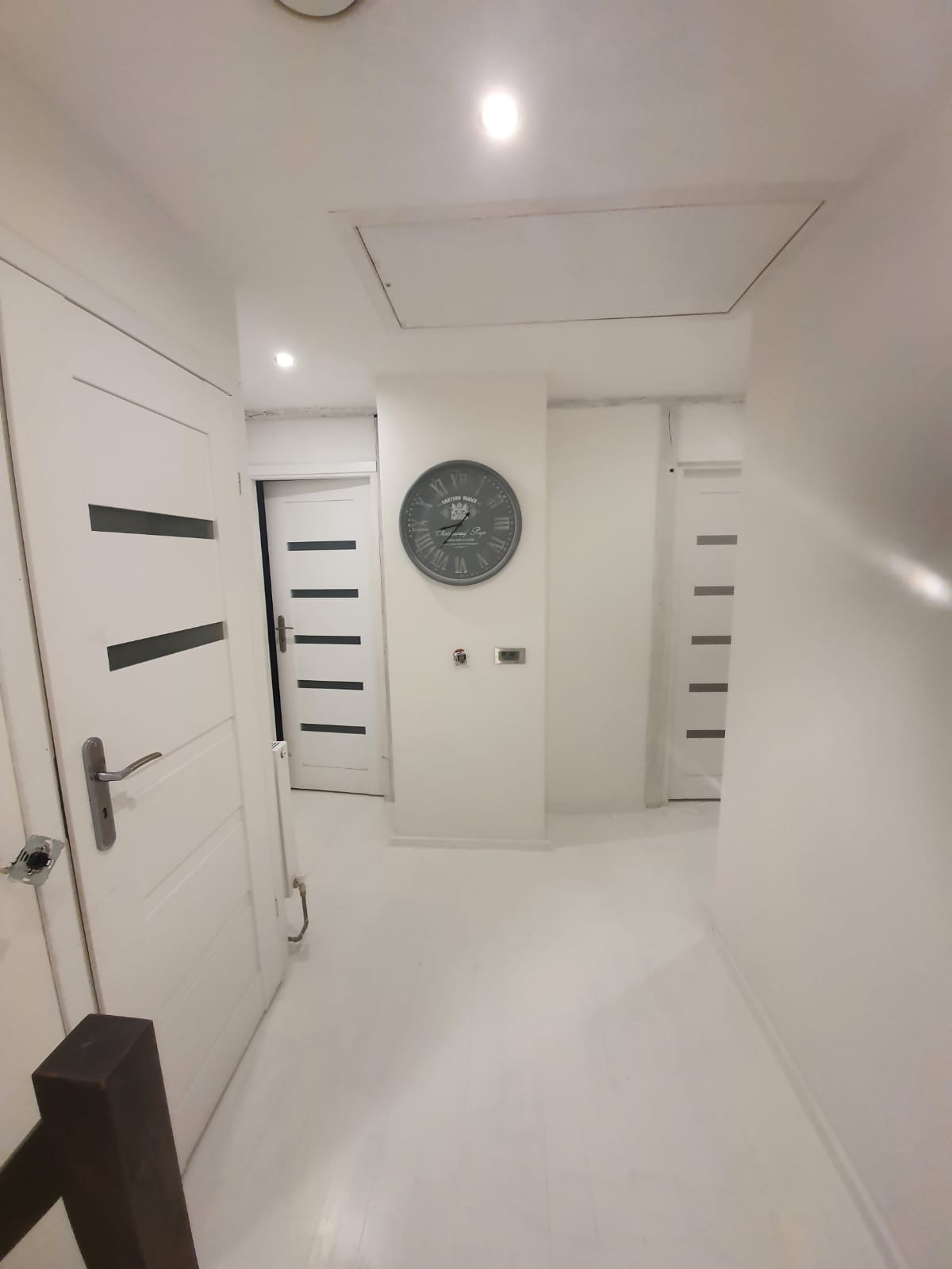 Jasny korytarz z białymi ścianami, drzwiami z poziomymi przeszkleniami i dużym zegarem na ścianie. Widoczne gniazdka elektryczne bez osprzętu.