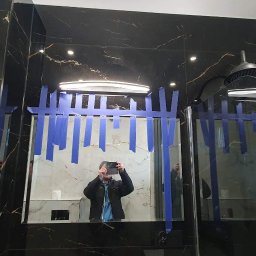 Łukasz Wentland - Nowoczesna łazienka z czarnymi i białymi marmurowymi płytkami, szklana kabina prysznicowa zabezpieczona niebieską taśmą malarską, widoczne odbicie osoby robiącej zdjęcie.