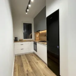 Nowoczesna kuchnia w wąskim pomieszczeniu: białe i szare szafki, drewniany blat, czarna lodówka, oświetlenie szynowe i panele podłogowe imitujące drewno.