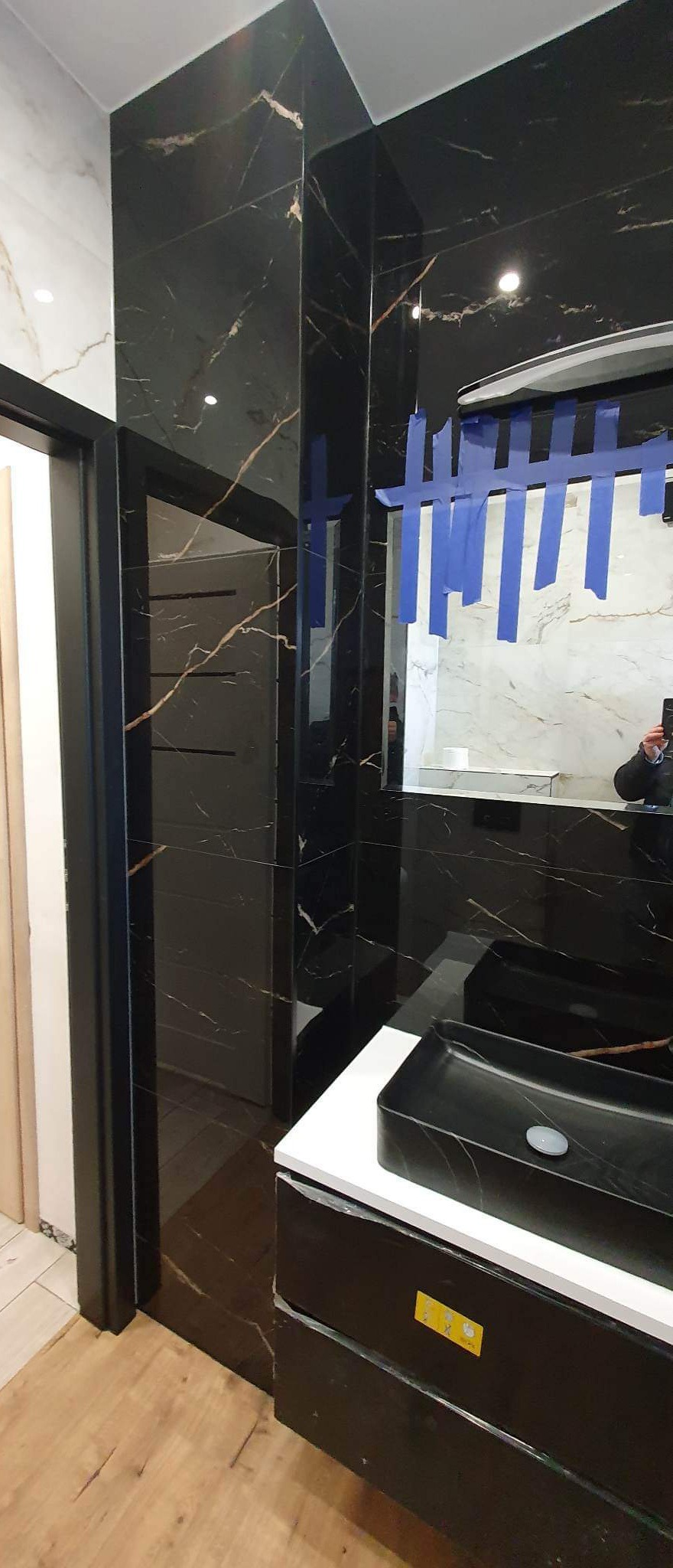 Wykończona łazienka z czarnymi płytkami imitującymi marmur z brązowymi żyłami, czarną umywalką na białym blacie i lustrem zabezpieczonym niebieską taśmą malarską.