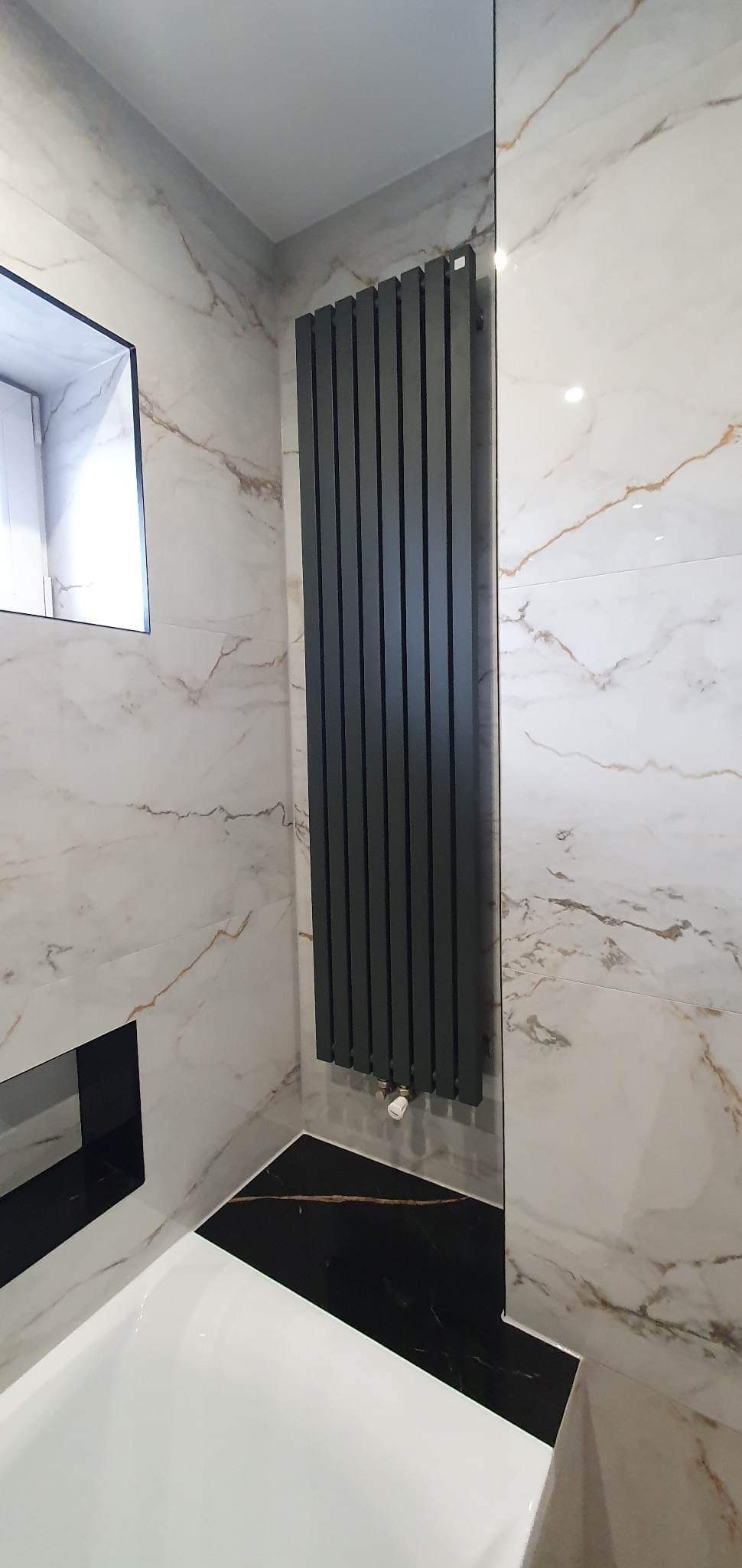 Nowoczesny, pionowy grzejnik panelowy w odcieniach szarości, zamontowany w łazience wykończonej białymi płytkami imitującymi marmur z delikatnymi złotymi żyłkami oraz czarnymi płytkami podłogowymi,...