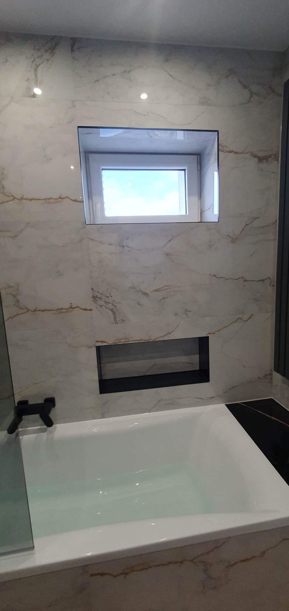 Biała wanna wypełniona wodą, otoczona marmurowymi płytkami z delikatnymi żyłkami, z wbudowanym oknem i półką w czarnych ramach.