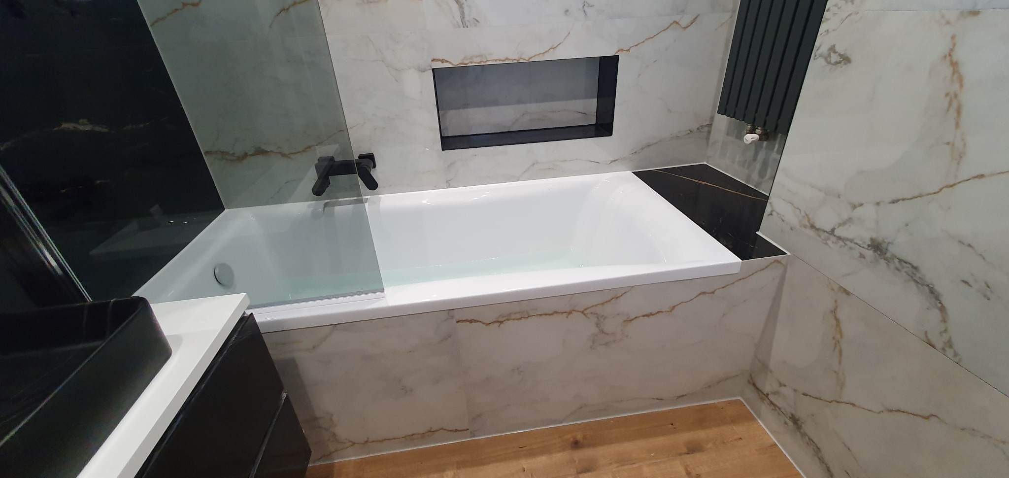 Wykończona łazienka z białą wanną obudowaną marmurowymi płytkami z brązowymi żyłami, czarna bateria umywalkowa, czarny grzejnik drabinkowy i wnęka w ścianie obłożona czarnymi płytkami.