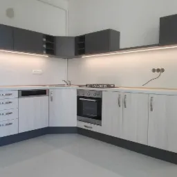 Kątowe zabudowa kuchenna z szarymi szafkami wiszącymi i frontami imitującymi jasne drewno, oświetlenie LED pod szafkami, widoczne gniazdka elektryczne wymagające wykończenia.