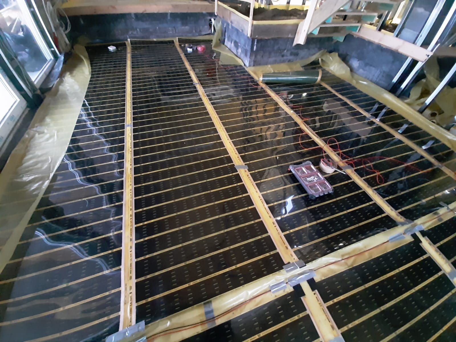 Montaż maty grzewczej na podłodze, widoczne czarne panele grzewcze z żółtymi paskami, zabezpieczone folią, z narzędziami i akcesoriami obok.