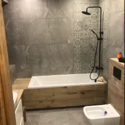 Nowoczesna łazienka z szarymi płytkami imitującymi marmur, dekoracyjnym pasem płytek wzorzystych, białą wanną obudowaną drewnopodobnymi panelami, czarną armaturą natryskową i podwieszanym bidetem.