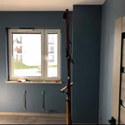 Świeżo pomalowane na niebiesko ściany pokoju z zamontowanym oknem i grzejnikiem, widoczna drabina malarska i fragment drewnianych drzwi.